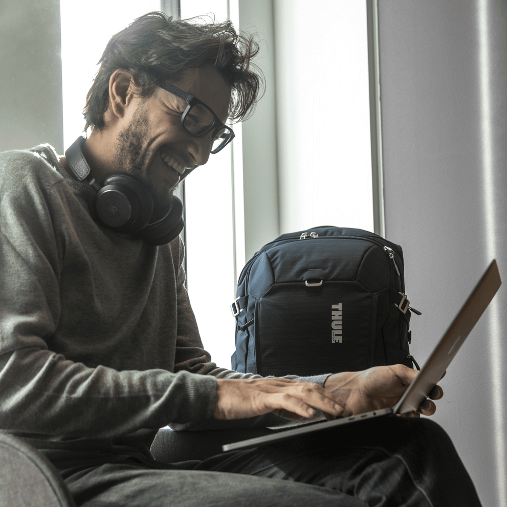 A man sits on a chair near a window, laughing at his laptop with a Thule Construct backpack in the background.