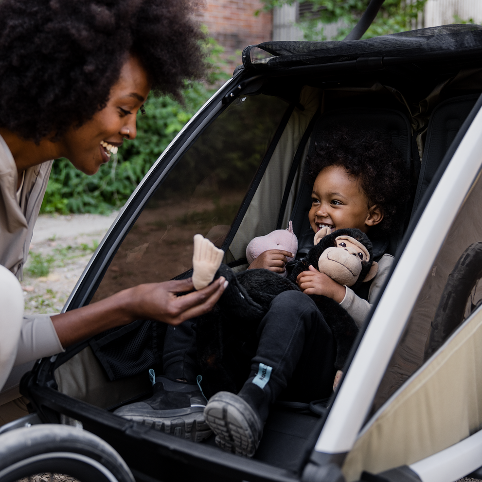Smiling child sitting inside a Thule Chariot Cross 2 bike trailer and stroller, while their parent leans in and smiles during a walk.