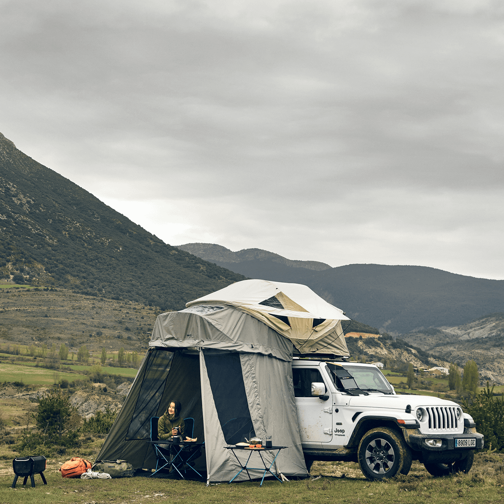 A car with a roof top tent that has an annex stands in front of a mountain