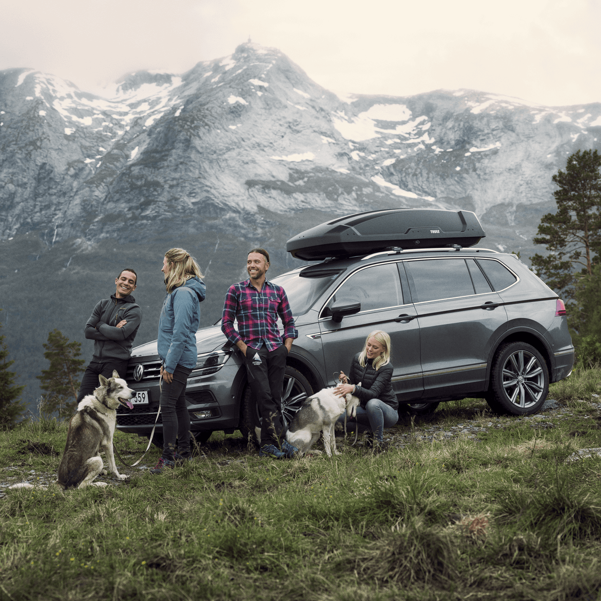 A group of people stands in front of a car with a roof box parked outdoors by a scenic mountain