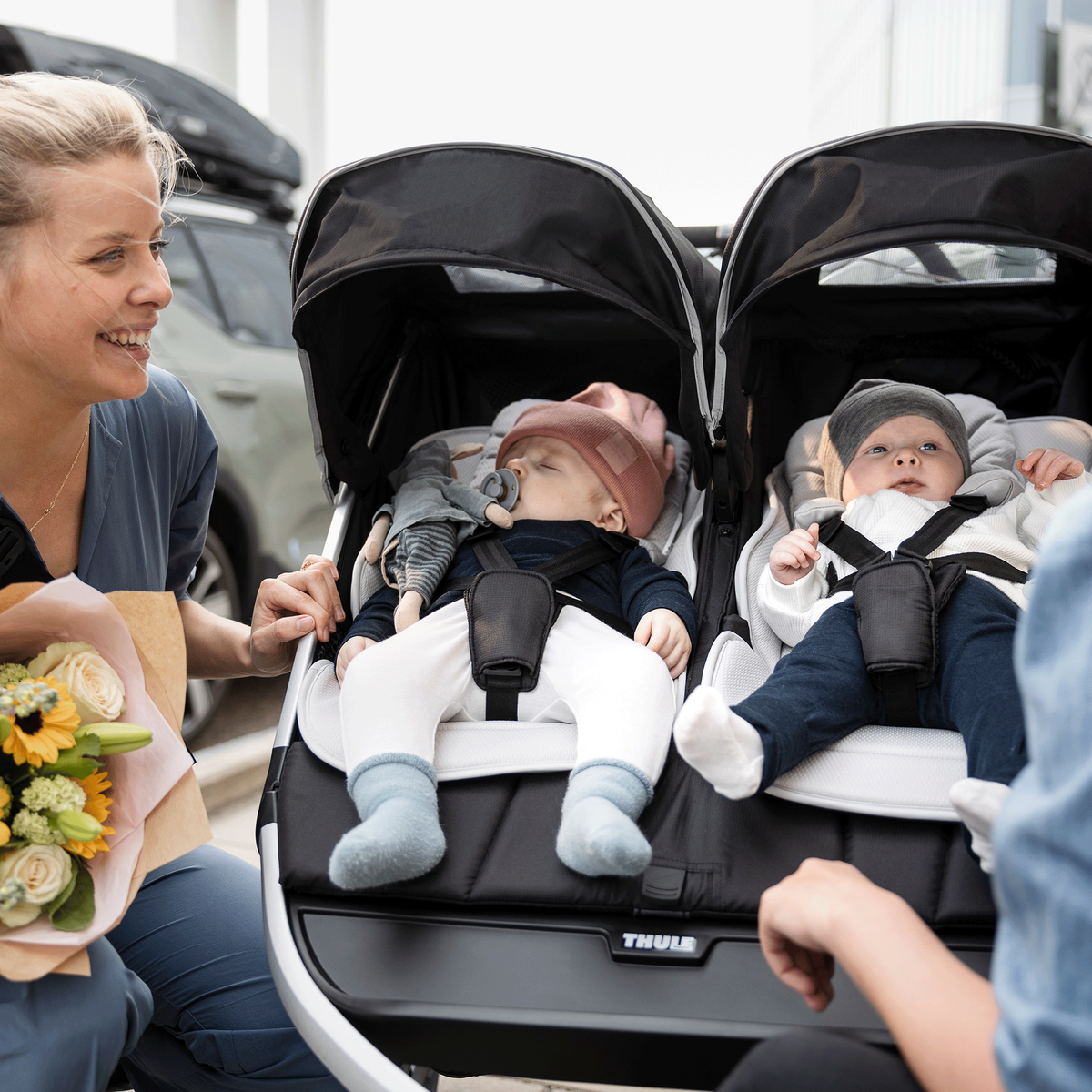 A woman holds a bouquet of flowers and tends to two kids in a black Thule Urban Glide 2 double jogging stroller.