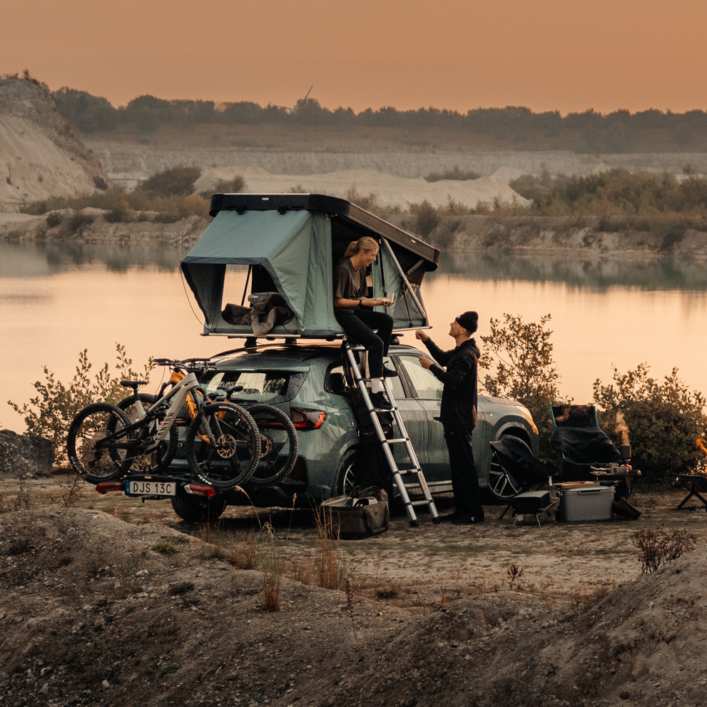Thule Widesky hardshell rooftop tent unfolded and mounted on the top of a car with two bikes on the towbar bike rack, parked in an outdoor setting and two people interacting by the car.
