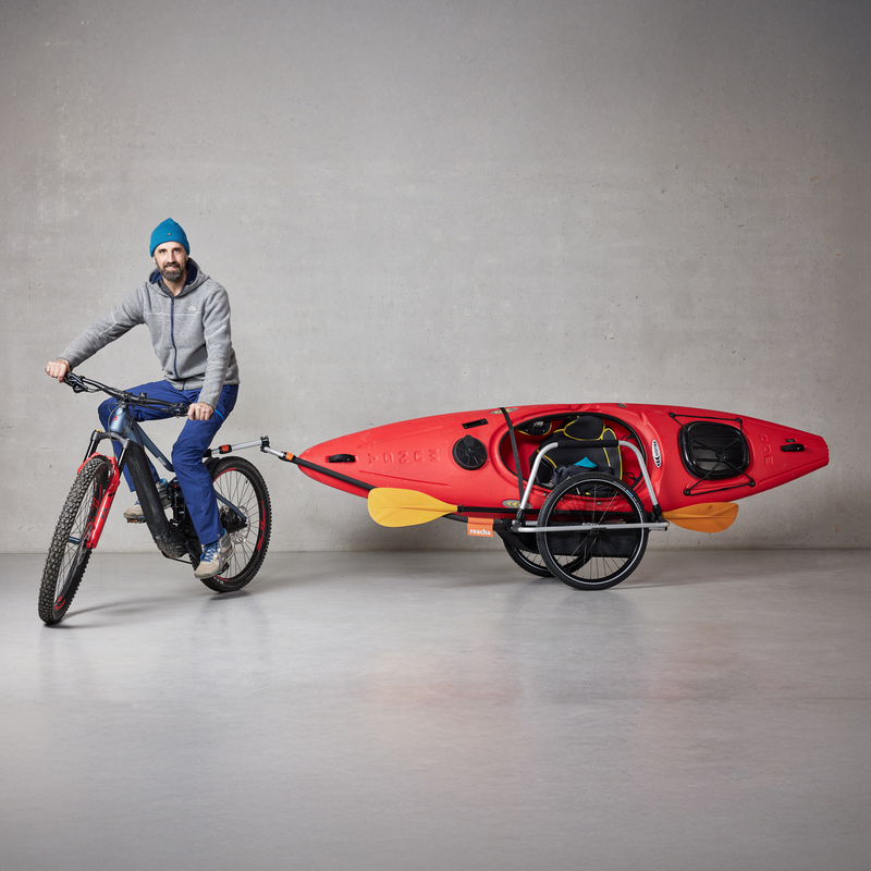 A man cycling indoors with a Reacha Sport bike trailer attached, carrying a bright orange kayak.