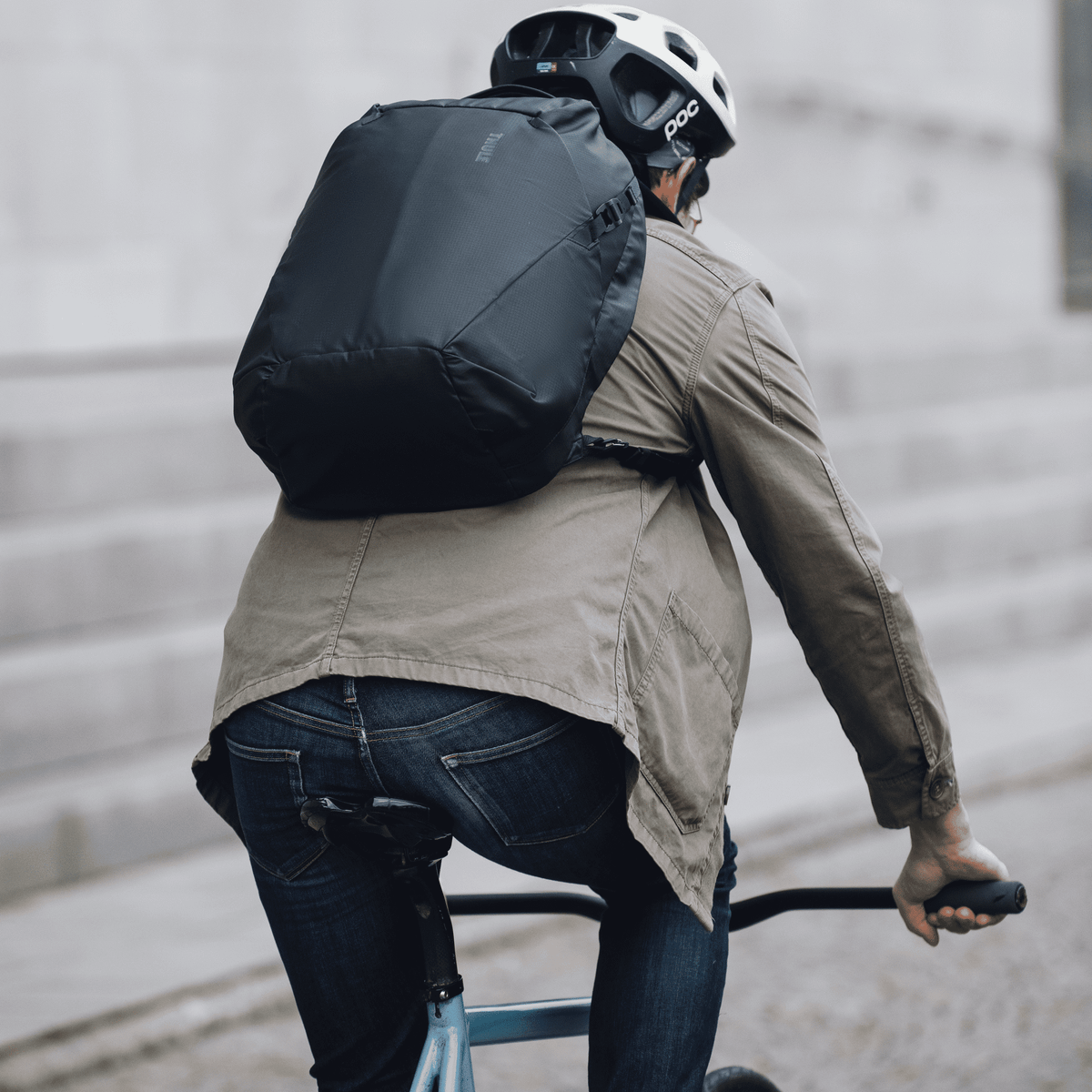 A man bikes down a street with a white helmet and a black Thule Tact backpack.