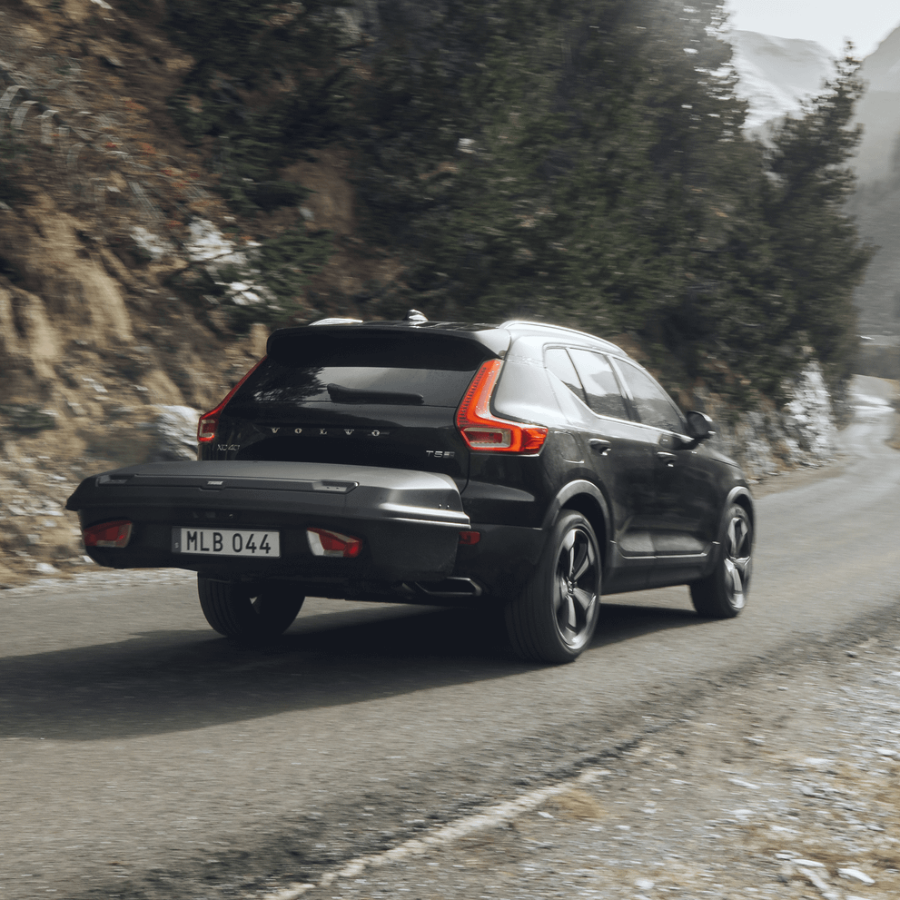 A black Volvo drives down a mountain street with a black Thule Arcos trunk cargo box
