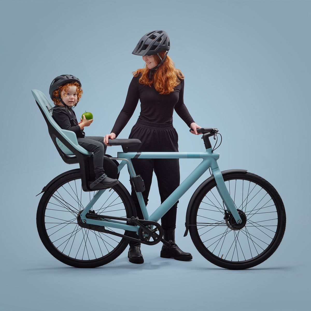A woman stands with a blue bike, blue background and her kid in a blue Thule Yepp 2 Maxi child bike seat.