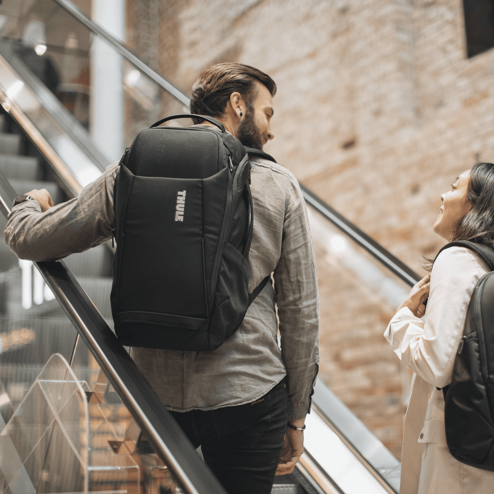 A man takes the elevator with a black Thule Accent Backpack 28L talking to a woman beside him.