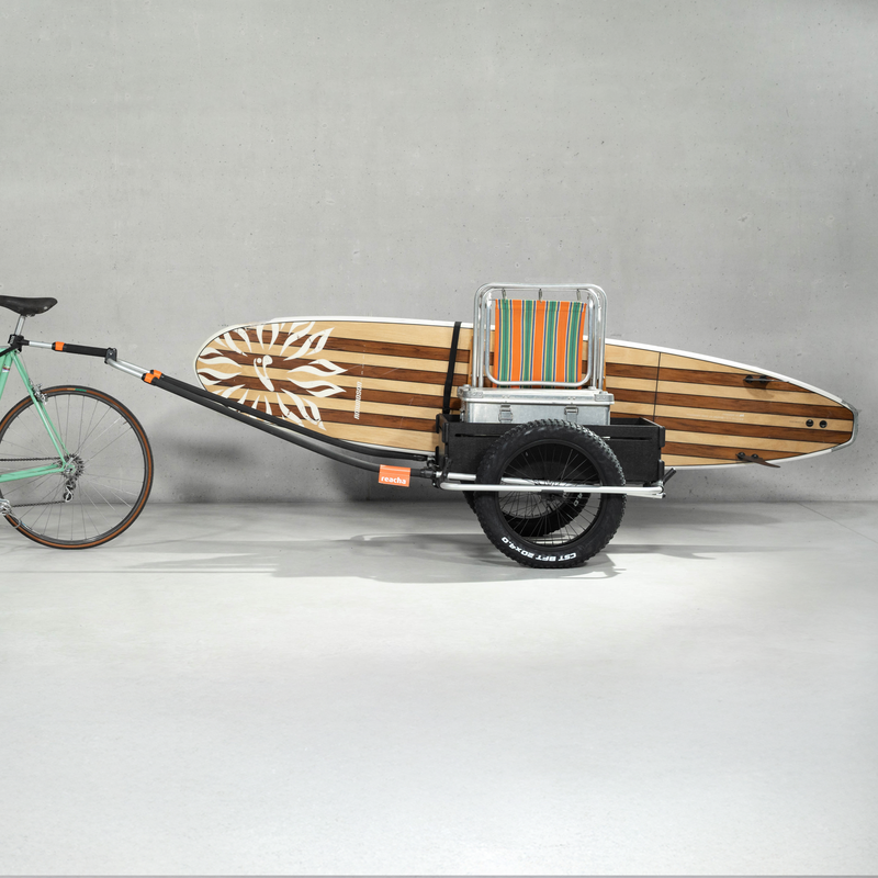 A green bicycle with a Reacha Sport Beach bike trailer attached, carrying a striped surfboard, displayed in a minimalist studio setting.