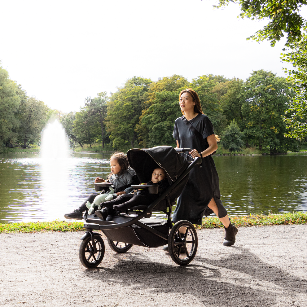 A woman walks down a gravel path in the park with her kids in a black Thule Urban Glide 3 double stroller.