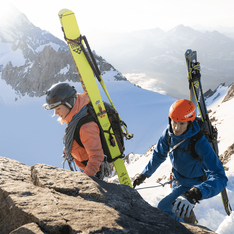 Two people climb up a snowy mountain with skis and rope attached to their Thule Stir Alpine 40L ski backpack.