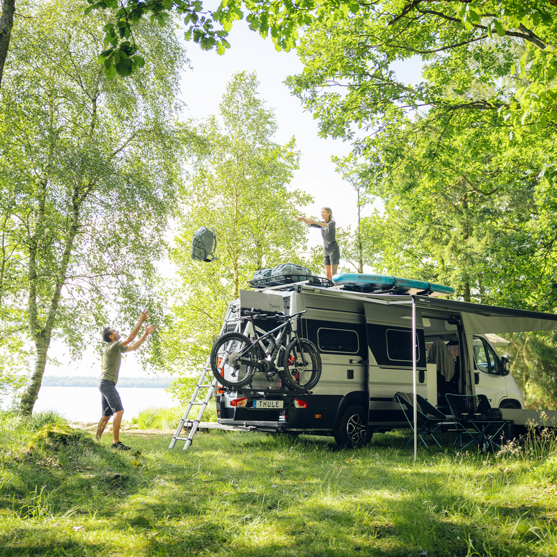 A camper van with the Thule VeloTrack bike rack and awning extended is set up in a lush, green campsite near a lake. A person is organizing gear, highlighting the ease of access and practicality of the setup.