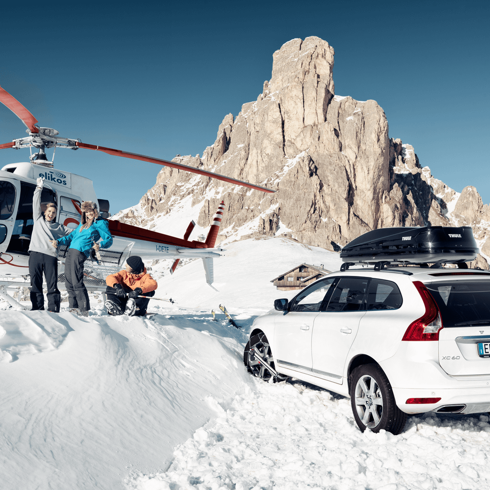 A group of people are standing on a snowy mountain between a helicopter and a car with a Thule Touring roof box.