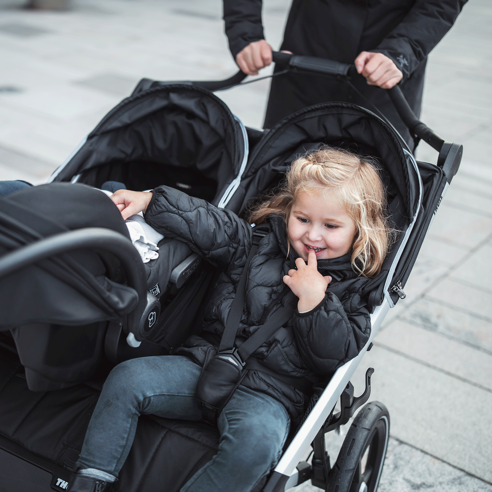 A toddler and a baby sit in the black Thule Urban Glide 2 double jogging stroller with a car seat attached.