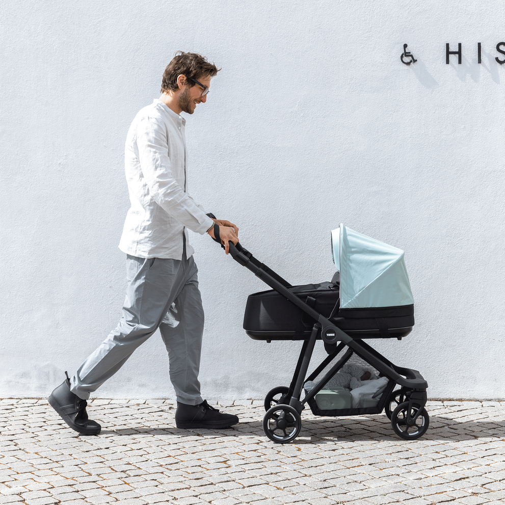 A man walks in front of a concrete wall with his blue Thule Shine bassinet stroller.