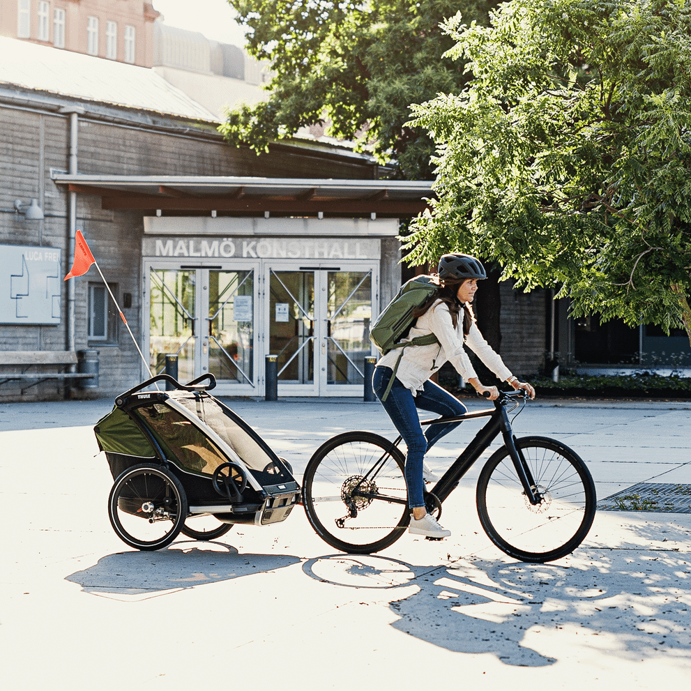 A woman with backpack and helmet bikes through the city with a Thule Chariot Cab cargo bike trailer.