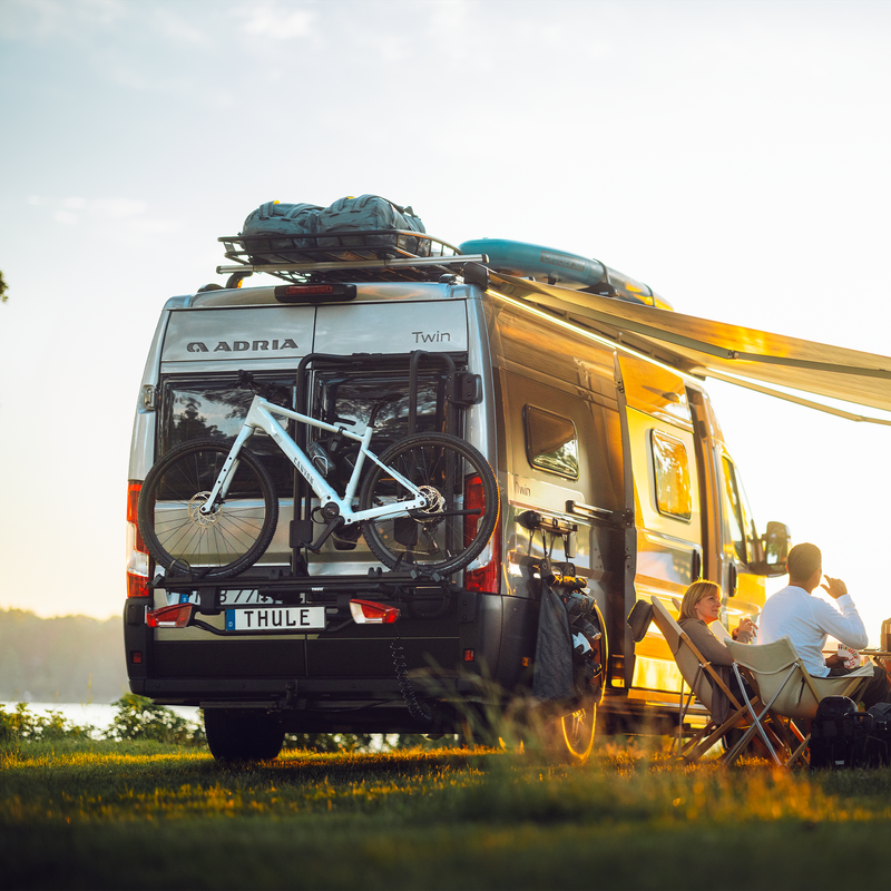 A camper van parked near a lake at sunset, featuring the Thule VeloTrack with the Thule Lightboard Van. The setup ensures bikes are mounted lower for better weight distribution and visibility of the van’s taillights.