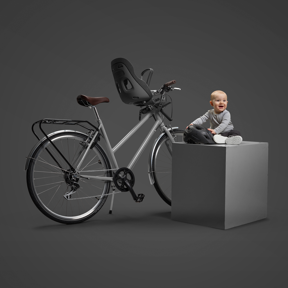 A bicycle is outfitted with a black child's bike seat mounted over the rear wheel, against a dark grey background. On the right, a toddler with a bright smile, wearing a grey long-sleeve shirt and black pants, sits on a white cube