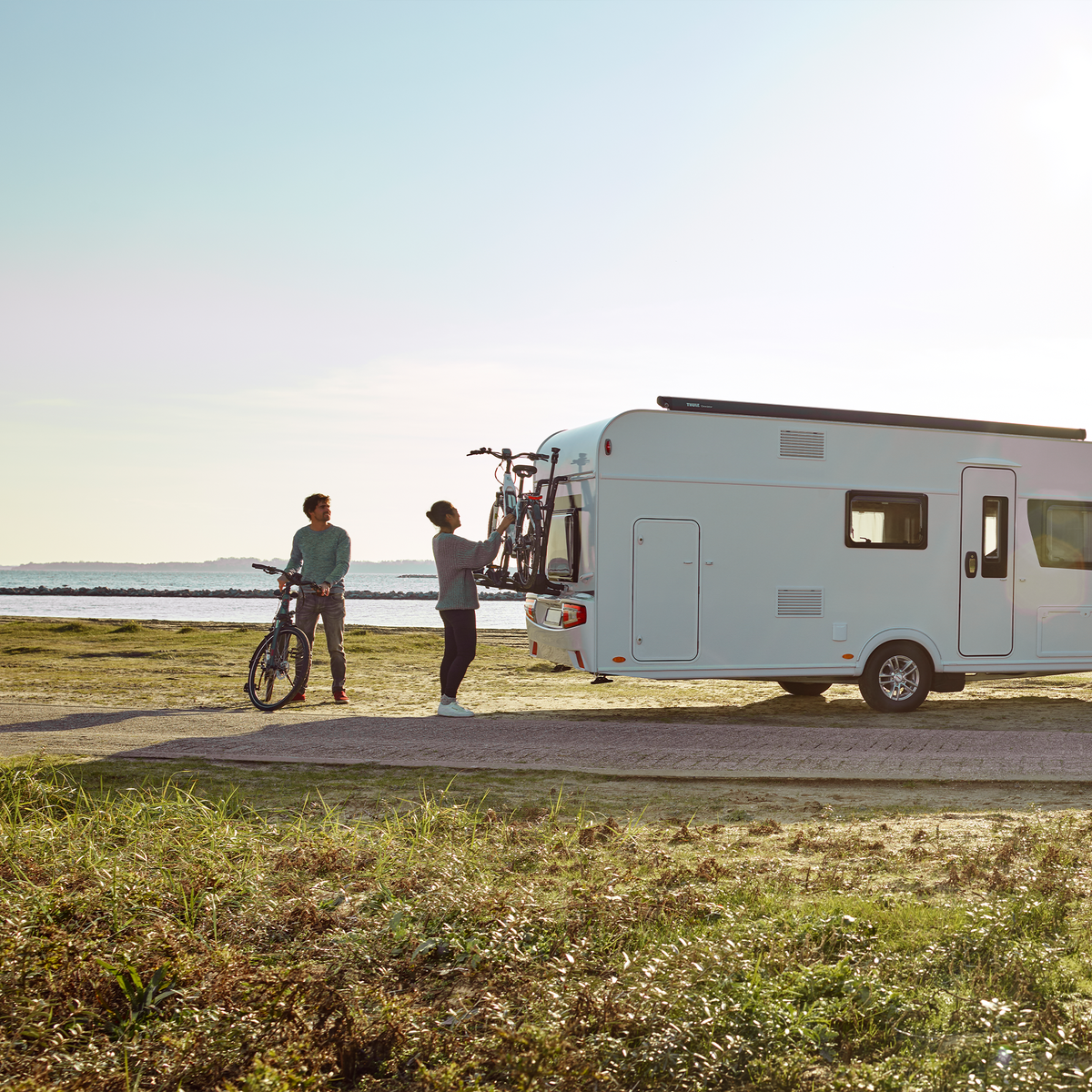 A white motorhome parked in a field with cyclists unloading bikes from a Thule Excellent Standard van bike rack.