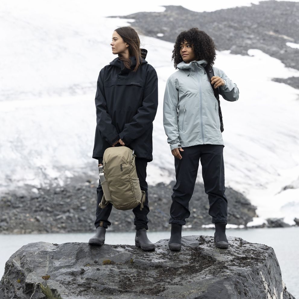 Two women stand on a stone next to a snowy mountain, holding Thule AllTrail backpacks.