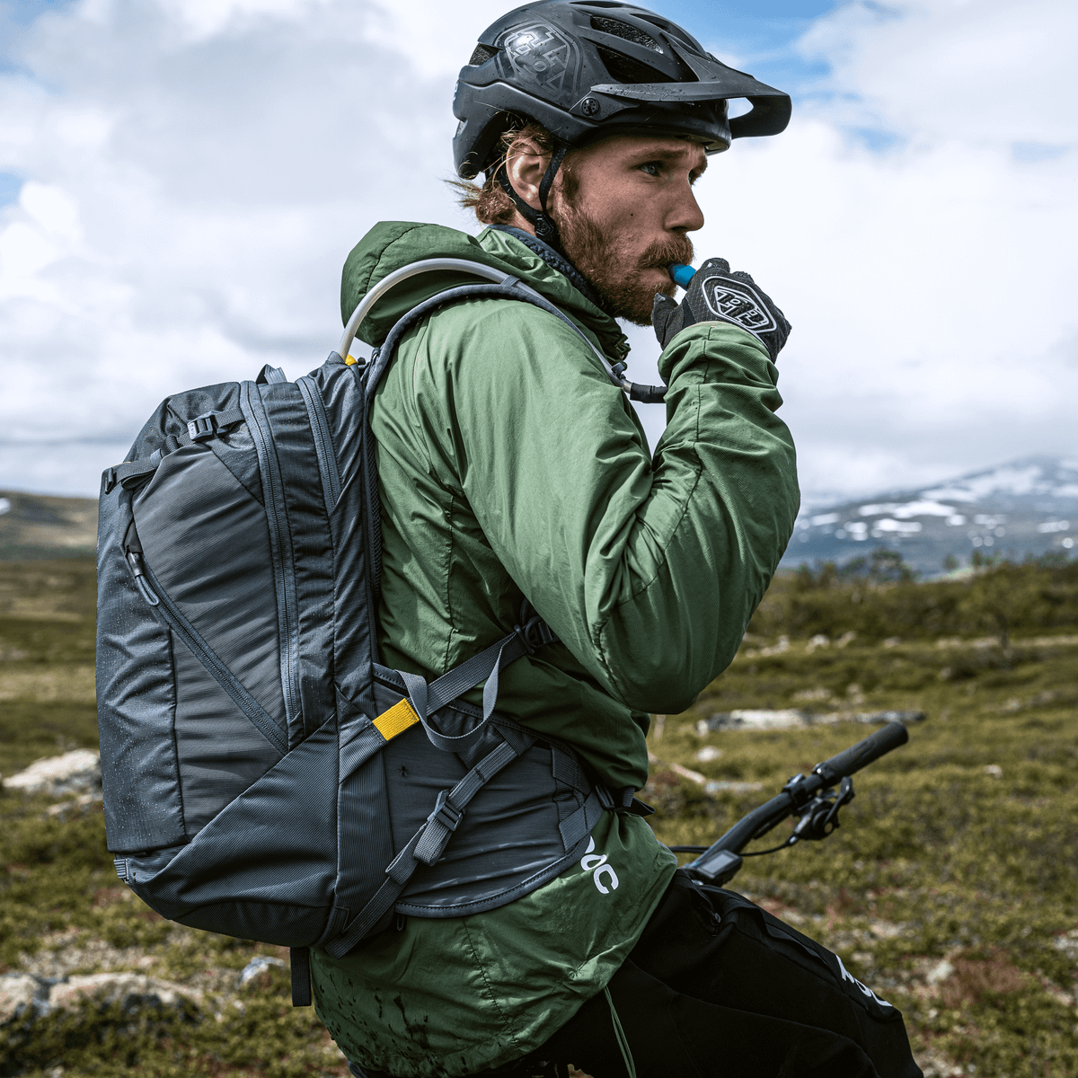 A mountain biker on a country trail is taking a sip from a blue Thule Rail 18L Hydration Backpack.