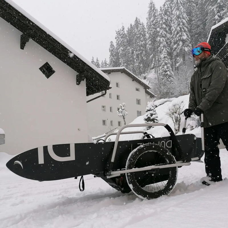 A man pulling a Reacha City bike trailer in snowy conditions, carrying a snowboard in a winter village setting.