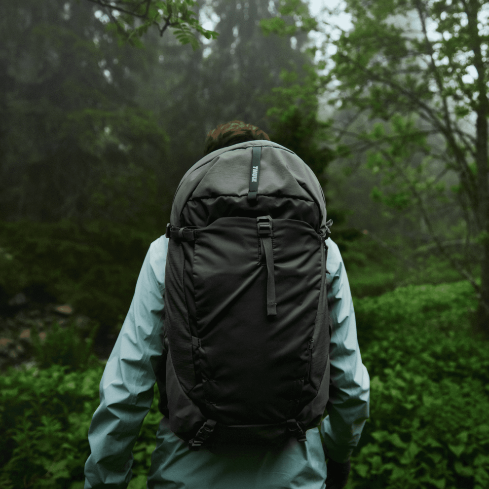 A person in a blue jacket walks through a misty forest carrying a black Thule Topio 30L hiking backpack.