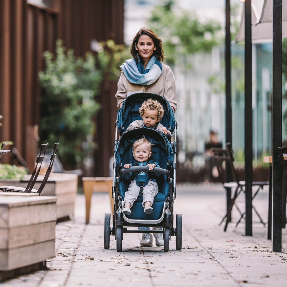On the sidewalk, a woman pushes her blue Thule Sleek double stroller with her two children.