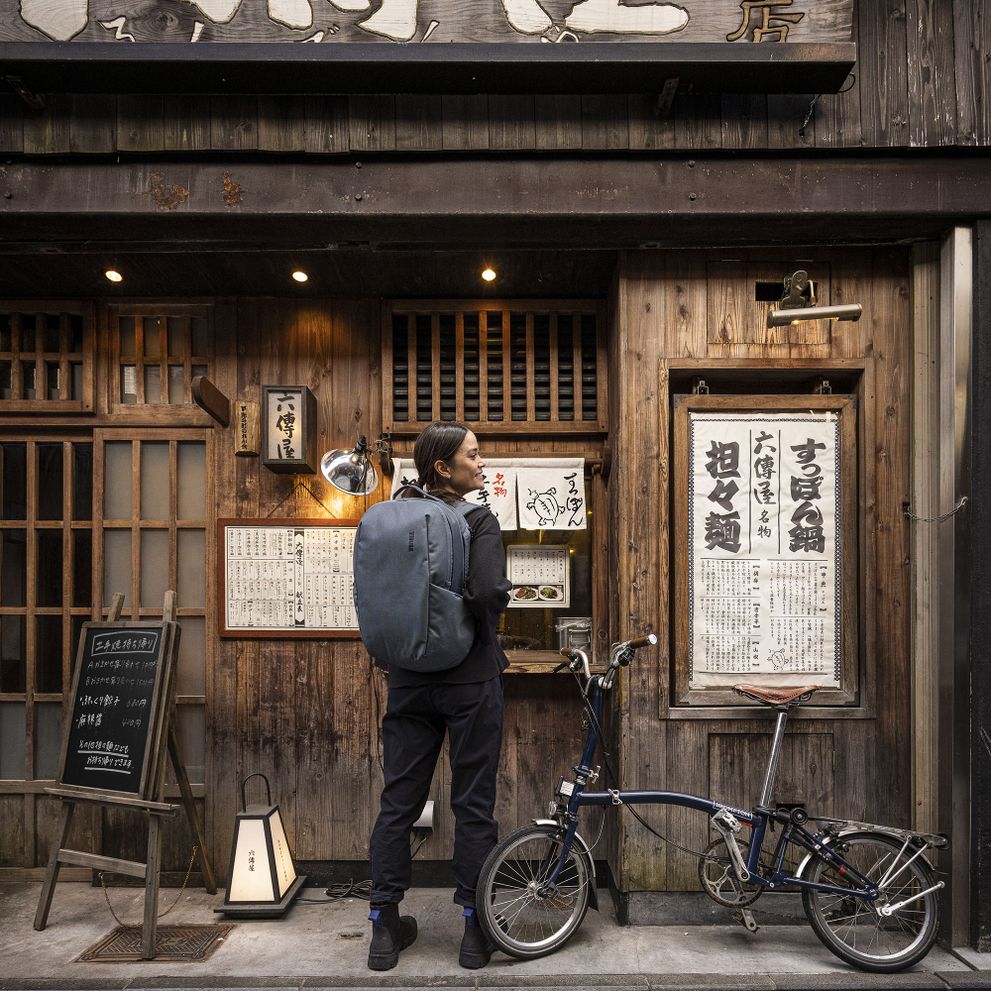 A woman stands outside of a wooden, Japanese shop with a bike and a blue Thule Subterra backpack.