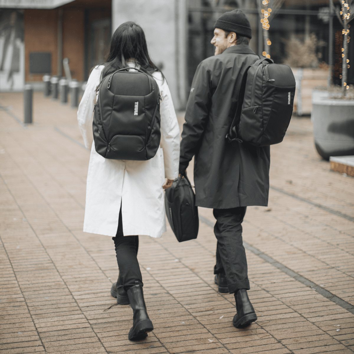 A man and a woman walk towards a glass building, chatting and carrying black Thule Accent backpacks.