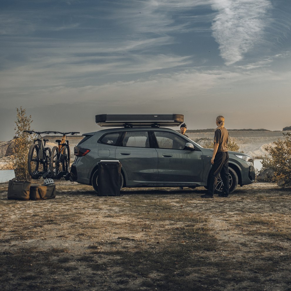 Thule Widesky hardshell rooftop tent unfolded and mounted on the top of a car with two bikes on the towbar bike rack, parked in an outdoor setting and two people interacting by the car.