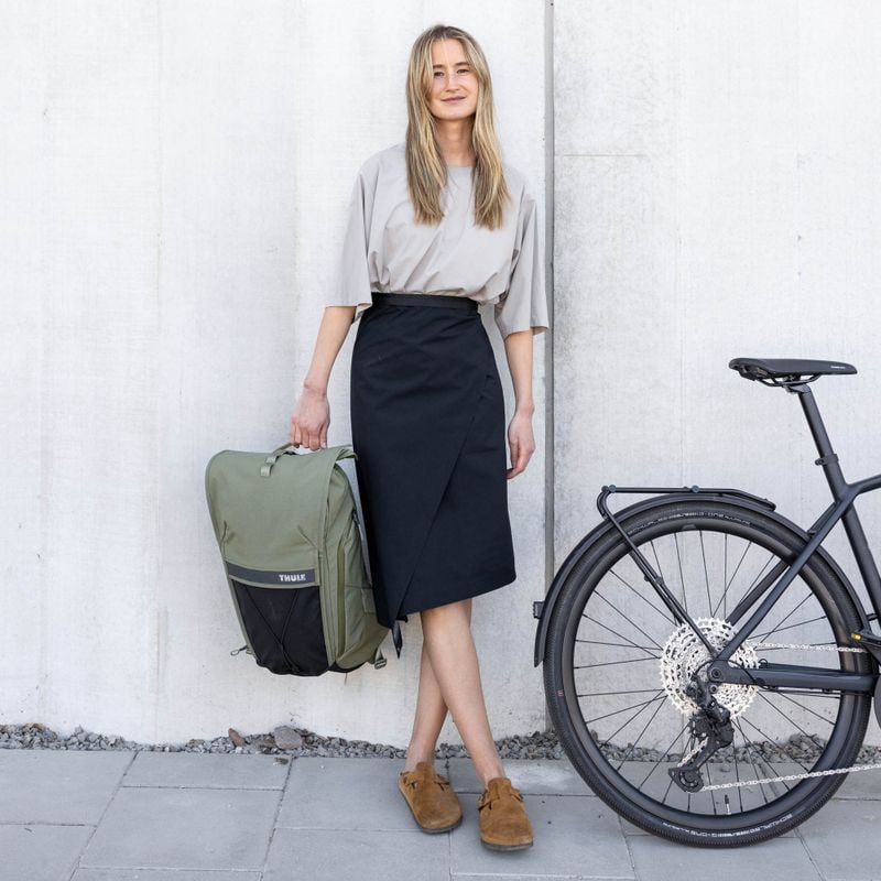 A woman with long blonde hair, wearing a beige top and black skirt, stands next to a bicycle holding a green Thule bag against a concrete wall.