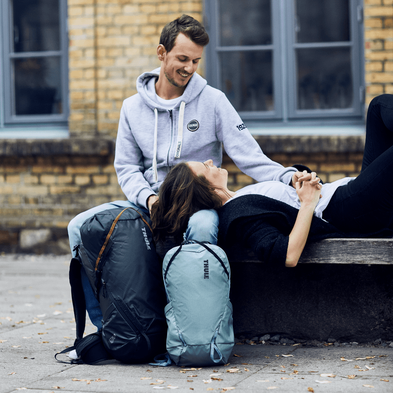 Two people stand and chat on a beach carrying yellow and blue Thule Stir 25L backpacks.