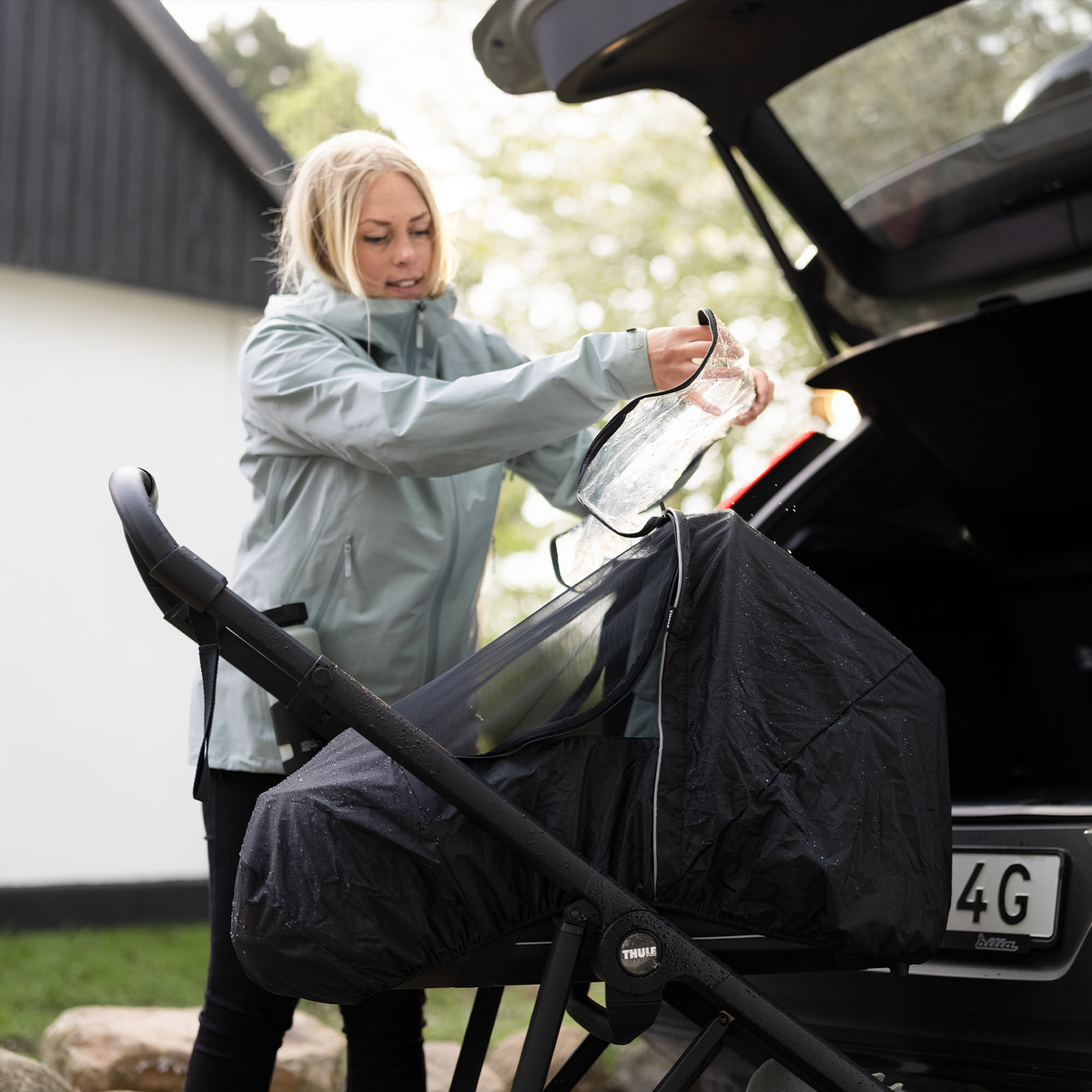 A woman opens the trunk of her car and assembles her stroller with a Thule Shine All-weather Cover.