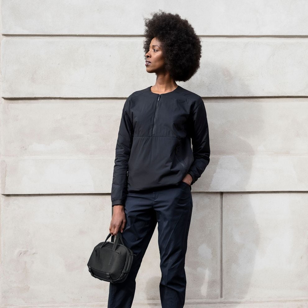 A woman wearing black stands against a brick wall holding a black Thule Subterra Powershuttle electronics organizer.