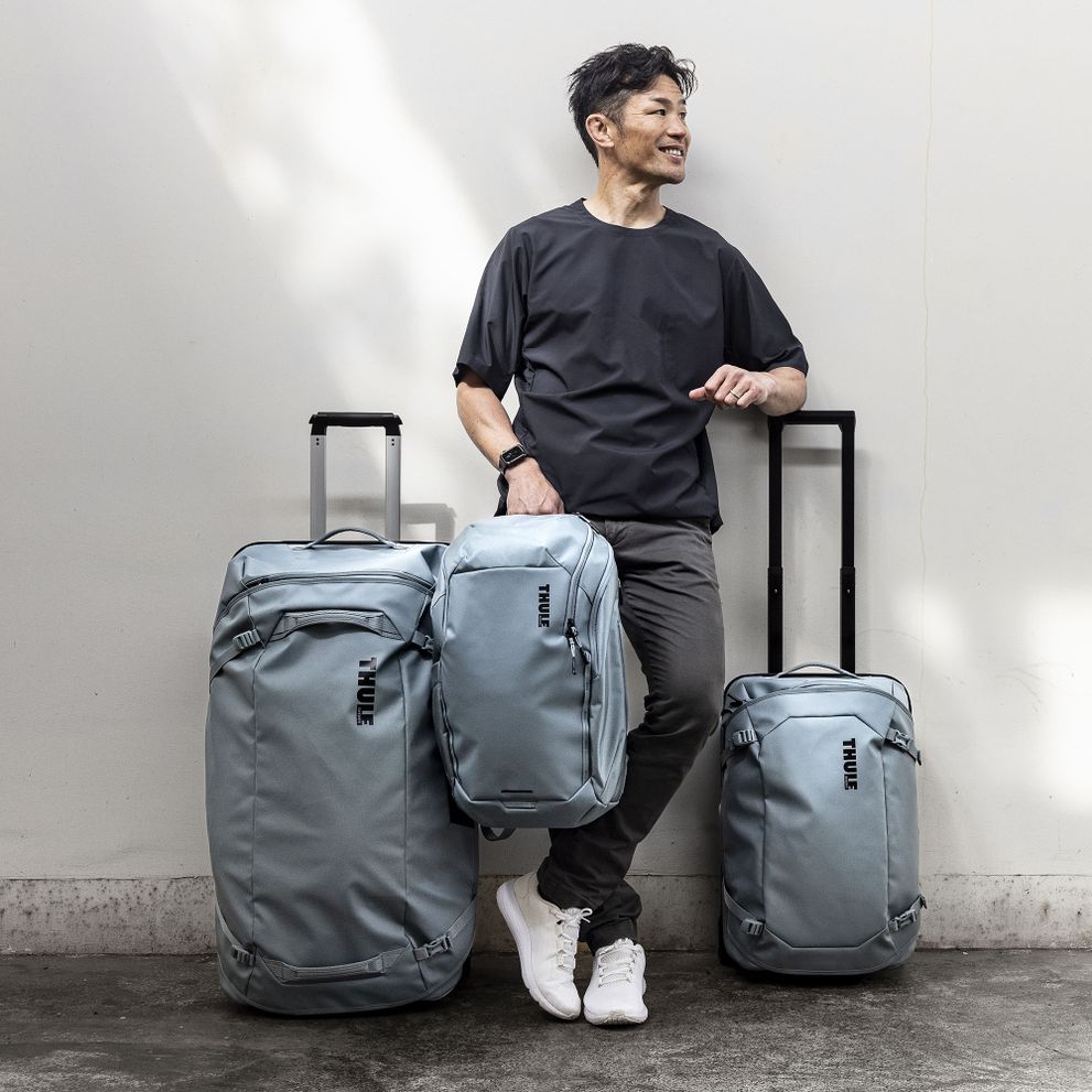 A man is leaning against a white wall with blue Thule Chasm suitcases and a Thule chasm backpack.