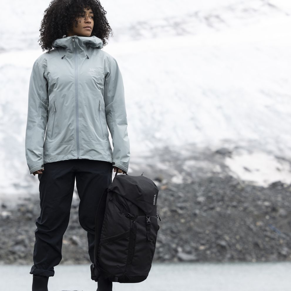 A woman stands beside a snowy landscape, wearing a blue rain jacket and holding a black Thule AllTrail backpack.