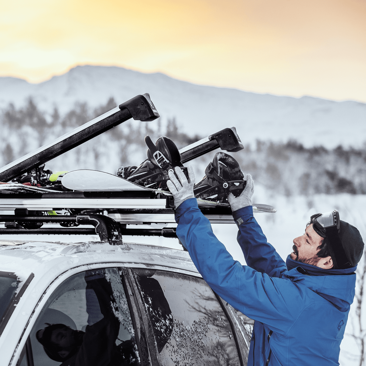 A man is fastening his snowboard on a car roof with a Thule SnowPack Extender