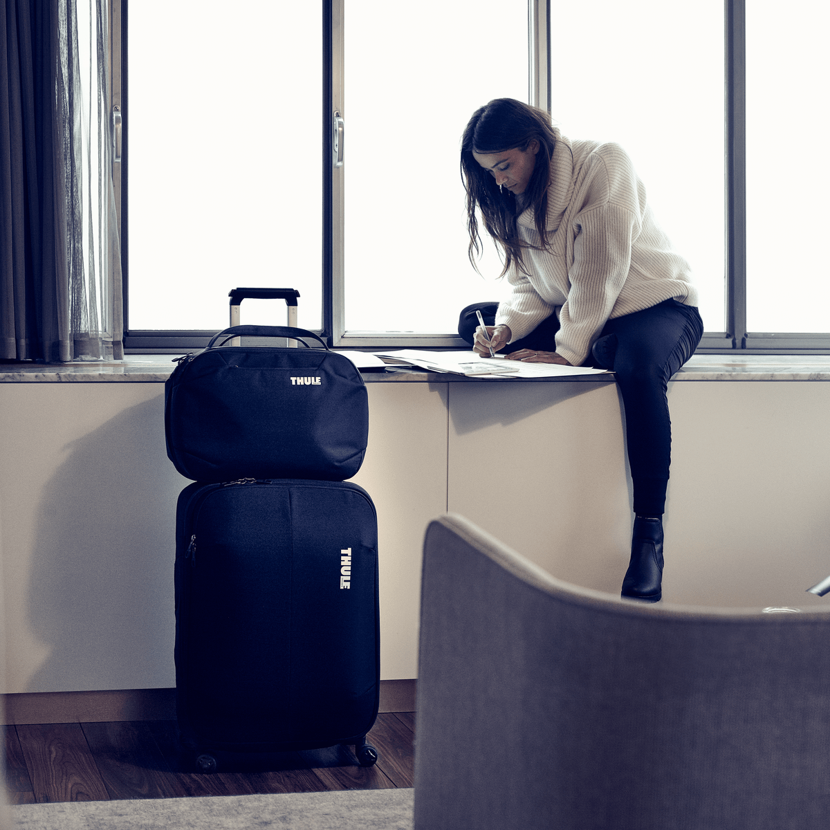 A woman sits on a window sill, with her backpack on a  on a Thule Subterra Carry-On Spinner suitcase.