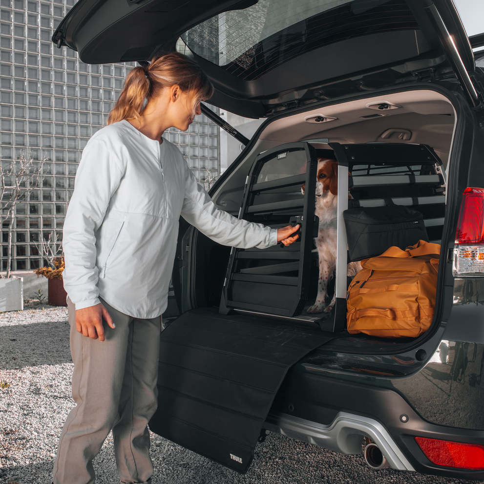 A woman shuts the door of the Thule Allax dog crate inside a car where a brown and white dog is sitting.