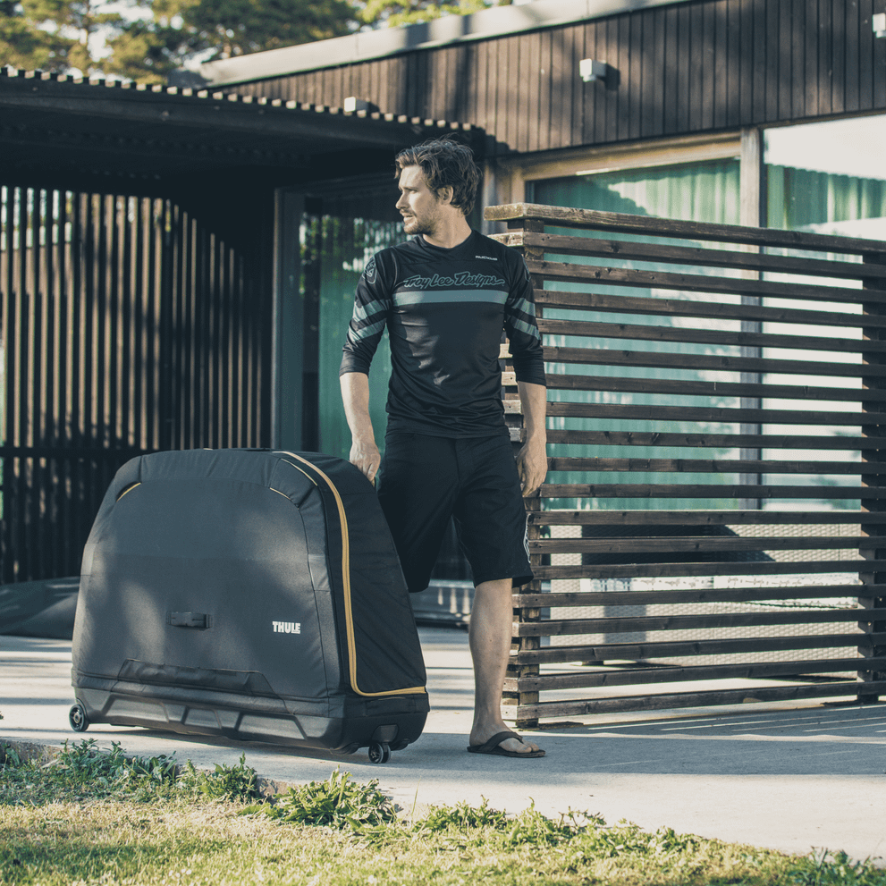A man stands outside a wooden building with a black Thule Roundtrip Road bike travel case.