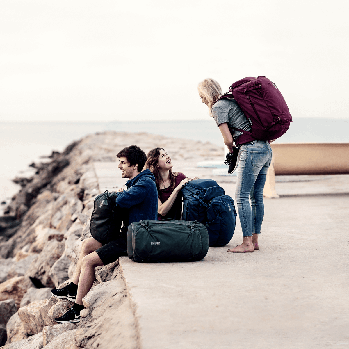 Three friends stand at a pier by the beach, all carrying Thule Landmark 70L travel backpacks.
