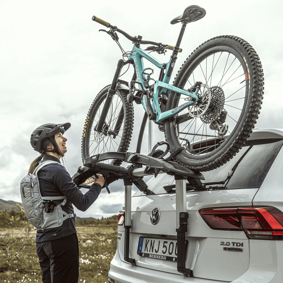 A cyclist loads her bike into a Thule Outway platform trunk bike rack in the countryside.