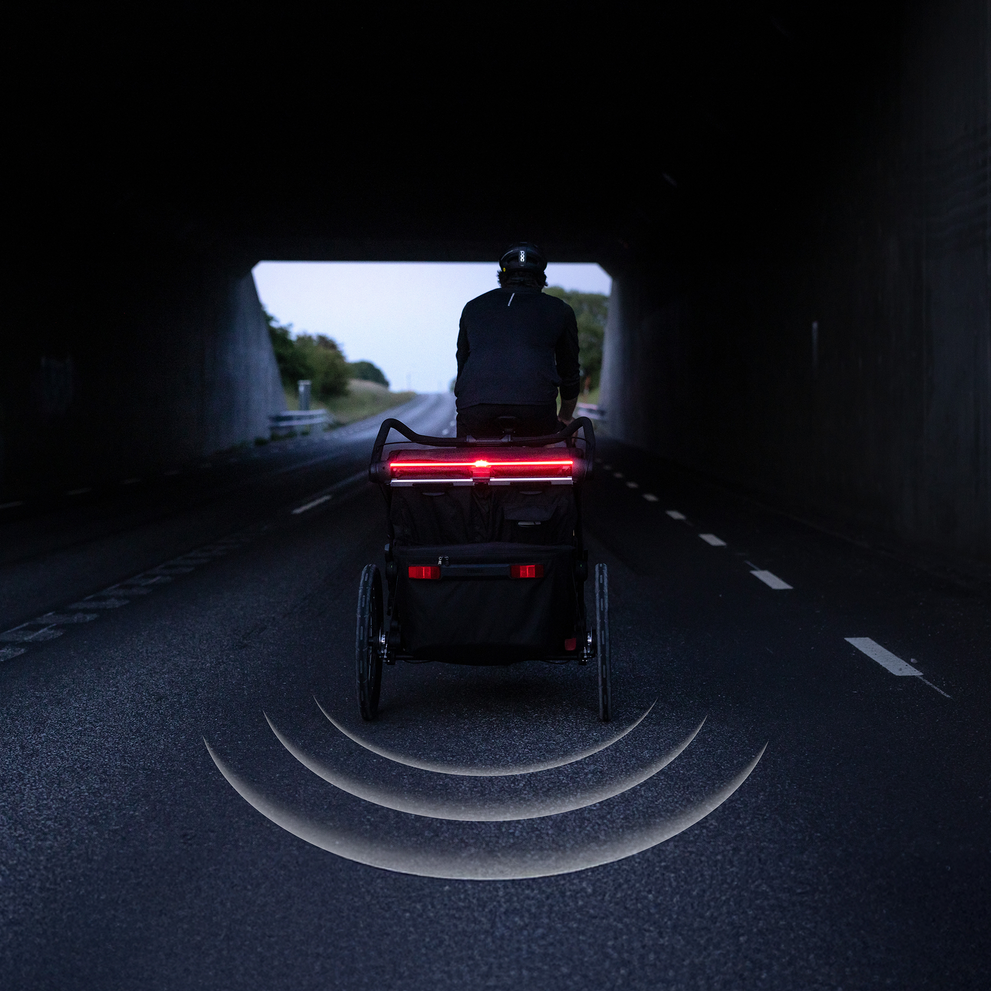Cyclist riding a bike with a trailer through a dark tunnel, featuring a bright red rear light mounted using a Thule mount for Garmin, with sensor-style light beams illustrated on the road behind.