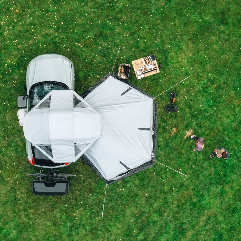An overhead view of Thule Approach 2 soft-shell rooftop tent unfolded and mounted with awning on the top of a car with one bike and Thule Santu cargo box on the towbar bike rack, parked in an outdoor setting and a family interacting by the car.
