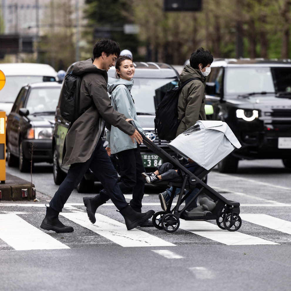 A couple walk across a crowded street with their toddler inside a Thule Shine Air Purifier canopy.