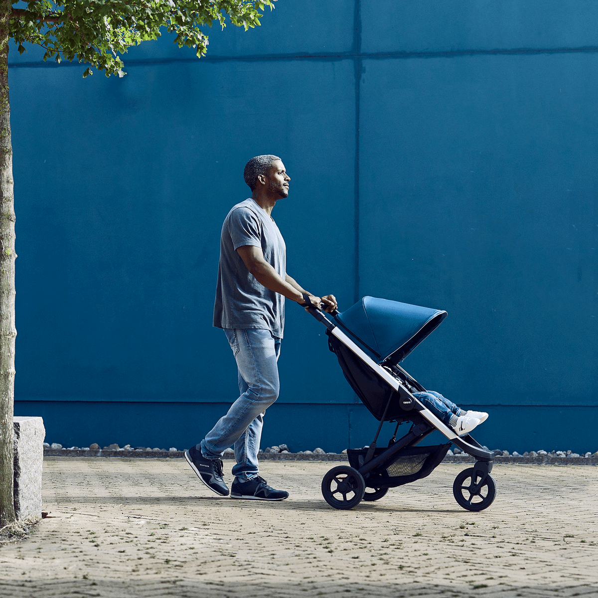 A man walks in front of a blue wall with a stroller that has a blue Thule Spring Canopy.