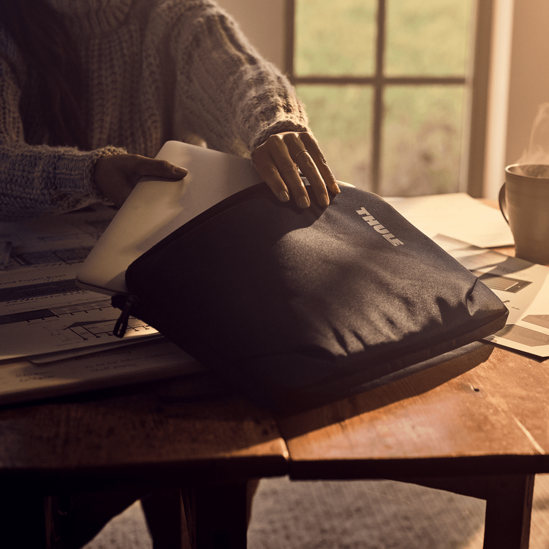 A woman puts tablets in a Thule Subterra MacBook Sleeve on a table with sunshine streaming in from the window.