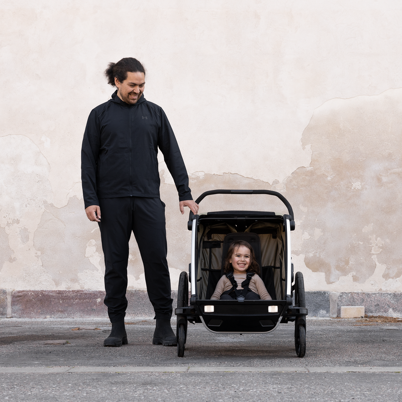 A man stands next to a brown Thule Chariot kids bike trailer wearing all black.