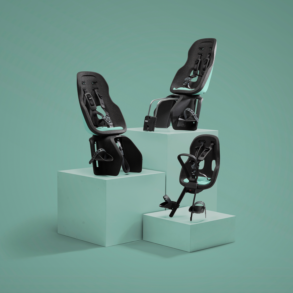 Tres asientos para bicicletas de niños se exhiben a diferentes alturas sobre pedestales blancos contra un fondo verde neutro. Cada asiento cuenta con un diseño elegante con un arnés de seguridad ajustable y correas para los pies