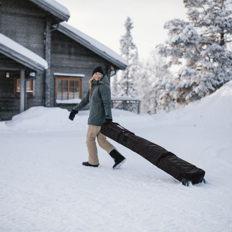 A woman stands in the snow outside a cottage wheeling her Thule RoundTrip travel snowboard bag.